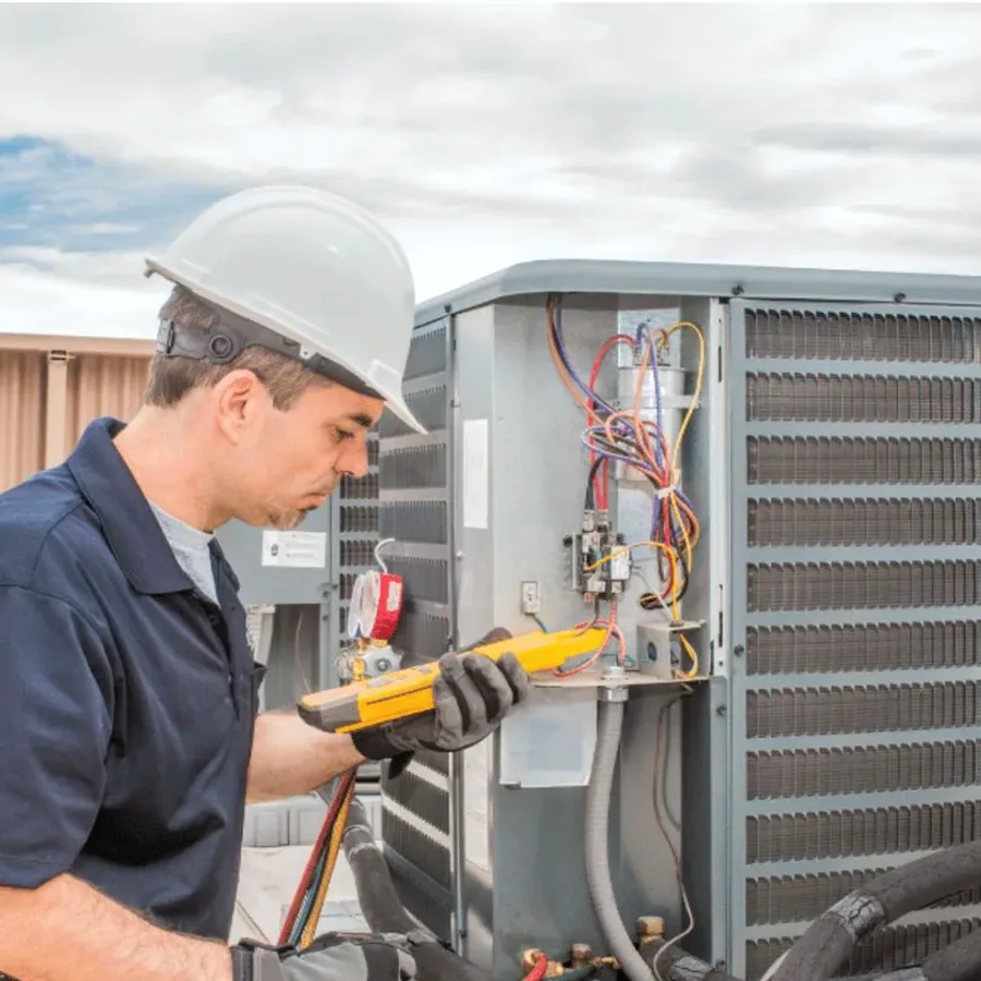 HVAC technician in a hard hat testing an outdoor air conditioner with a multimeter